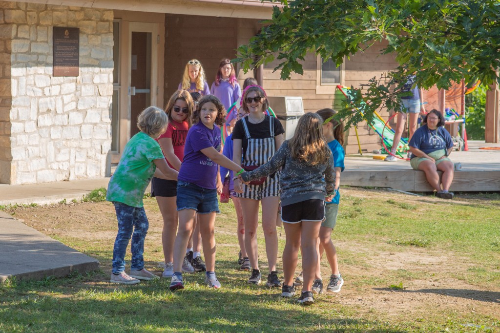 A group of young girls hold hands in a circle around their camp councelor, a young woman in striped overalls and a pink bandana. 