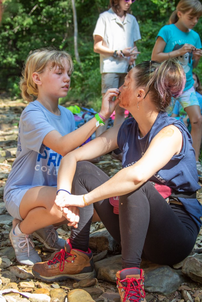 A young girl uses rock face paint on her counselor who sits on the rocky creek bed.  
